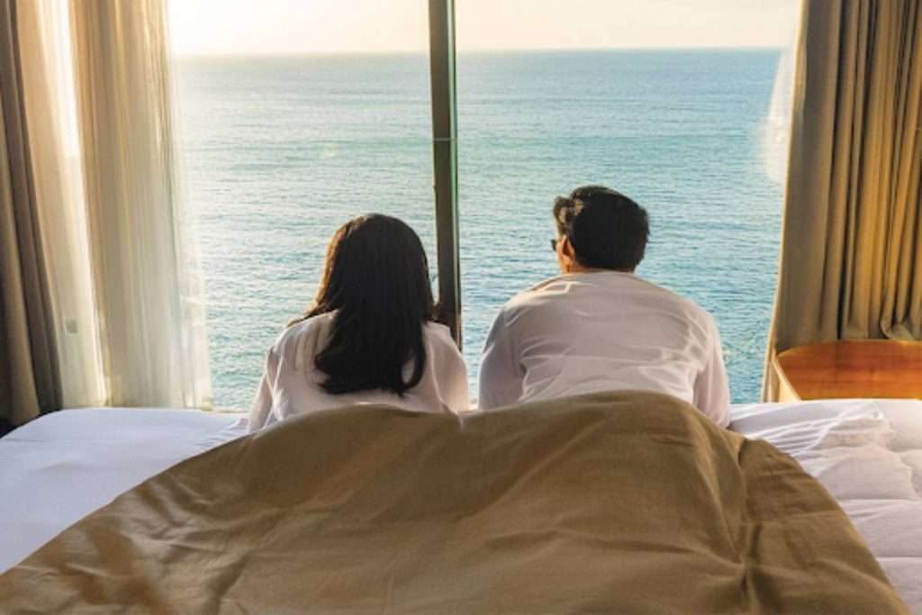A couple wearing white shirts lounges on a bed, gazing at the ocean through a large window