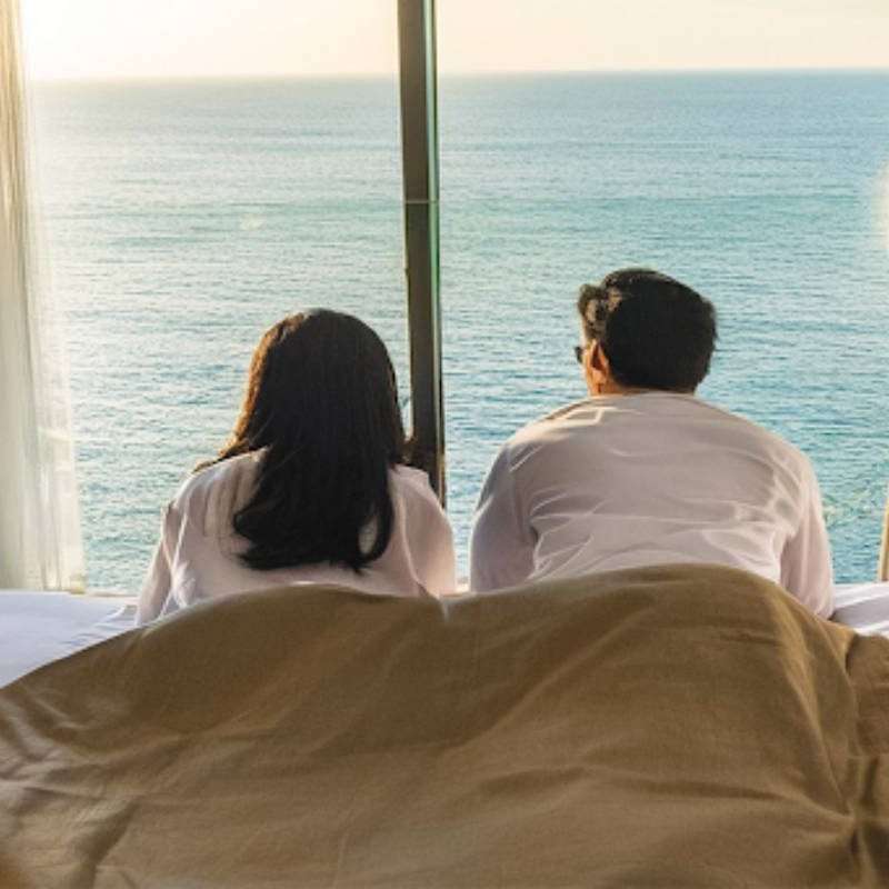 A couple wearing white shirts lounges on a bed, gazing at the ocean through a large window