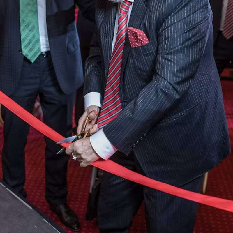 A man in a pinstriped suit cuts a red ribbon with scissors, surrounded by other suited individuals, suggesting a formal event