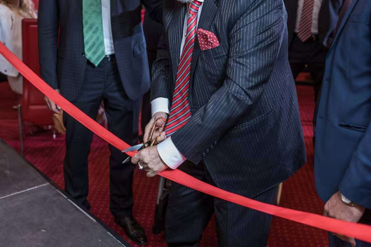 A man in a pinstriped suit cuts a red ribbon with scissors, surrounded by other suited individuals, suggesting a formal event